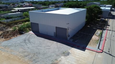 Vista aérea de galpão com fechamento metálico claro e duas portas de enrolar na fachada frontal, com pátio de cascalho à frente.